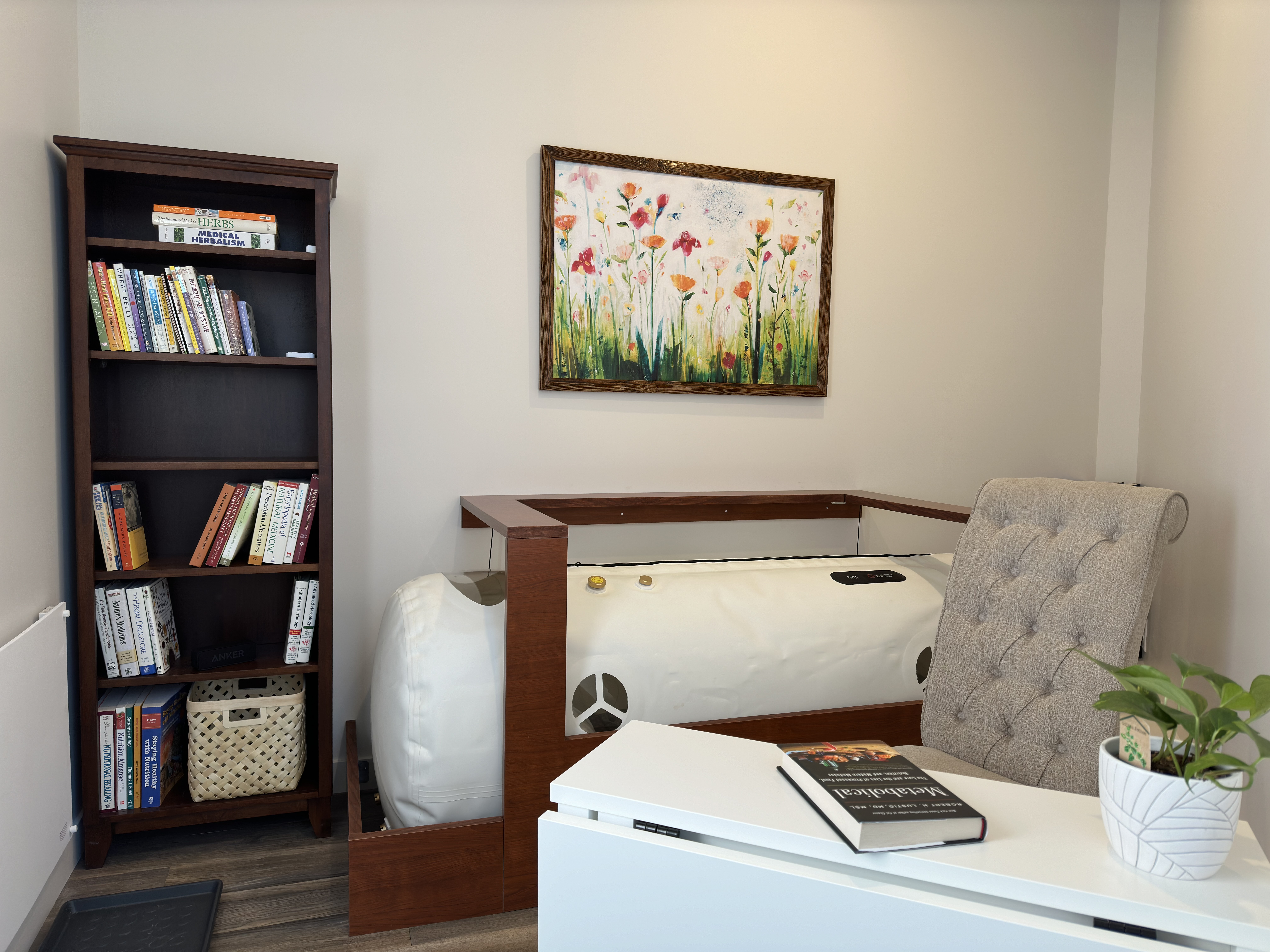Wide view of the calm relaxation room: wellness bookshelf, floral painting, soft-shell chamber and a tufted reading chair.